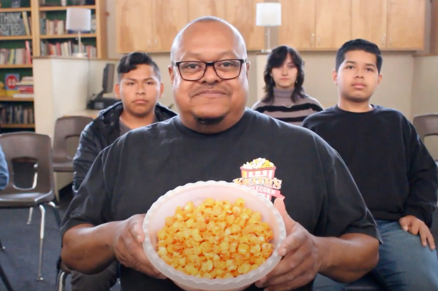 In this image, Keith Jackson, the founder of Keith's Kettle Corn, proudly presents a bowl of bright yellow popcorn at a school setting. He stands smiling at the camera, wearing a black T-shirt with the Keith's Kettle Corn logo, while three students in black attire observe from the background. The setting suggests a casual environment, possibly during a Popcorn Fundraiser event where Keith is interacting with students and discussing the importance of community engagement through such fundraising efforts.